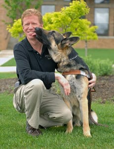 Keith kneeling outdoors on grass next to a German shepherd in harness licking his face
