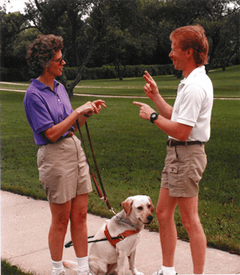 Keith as a younger man talking with a woman outside on a sidewalk. They are wearing khaki shorts and polos. A yellow lab in harness sits at the woman's side.