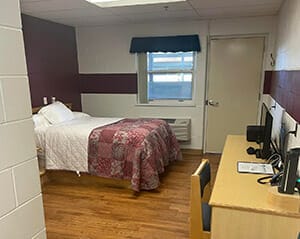 Interior of client room on campus with bed on left, desk on right, outside door in the background next to a window.