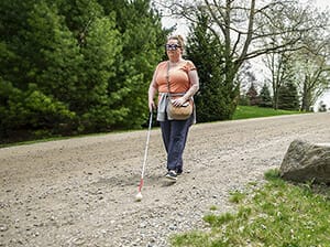 Woman with white cane walks on dirt road 