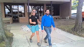 Screenshot of a woman walking next to a man in sunglasses on a sidewalk. The woman is holding the man's elbow and he is walking slightly ahead of her.