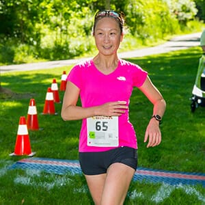 Woman in pink running shirt and black shorts with Bark & Brew 5K race bib on shirt crossing finish line on grass with trees in the background