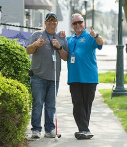 Two men smiling on sidewalk, one holding long white cane