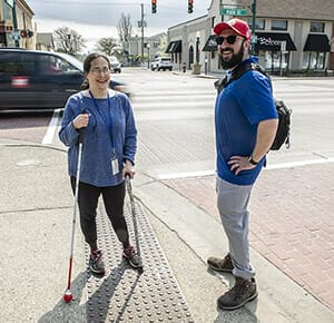 Woman with long white cane and man smiling standing near street curb