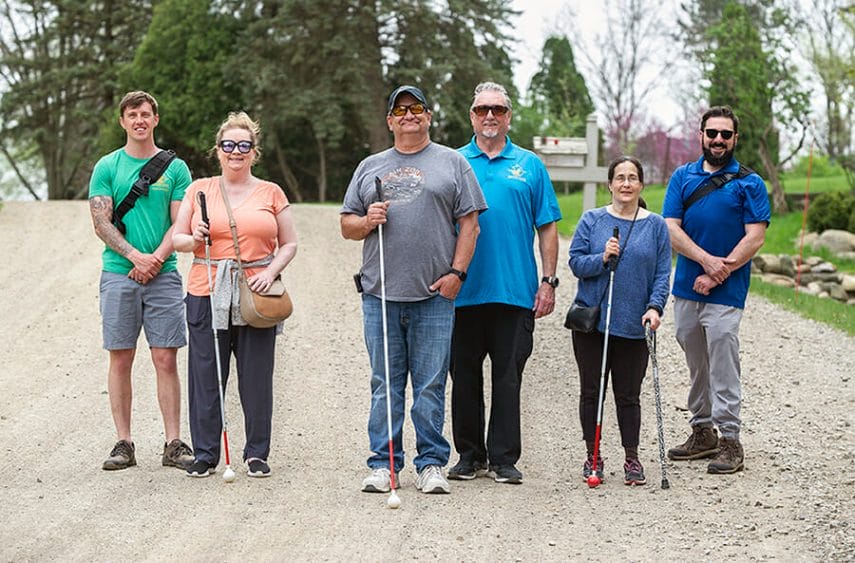 Six people, two women and four men, stand on a gravel road, smiling at the camera. Three people hold white canes.