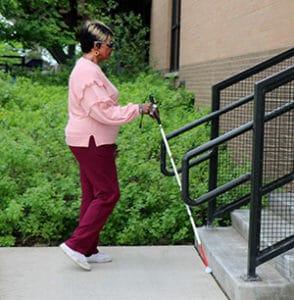 Woman with white cane walking toward outdoor concrete steps