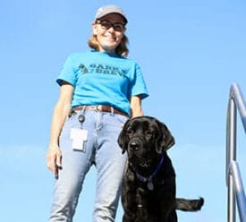 Smiling woman in blue t-shirt with Bark & Brew logo and jeans standing next to black lab with blue sky in background