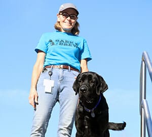 Smiling woman in blue t-shirt with Bark & Brew logo and jeans standing next to black lab with blue sky in background