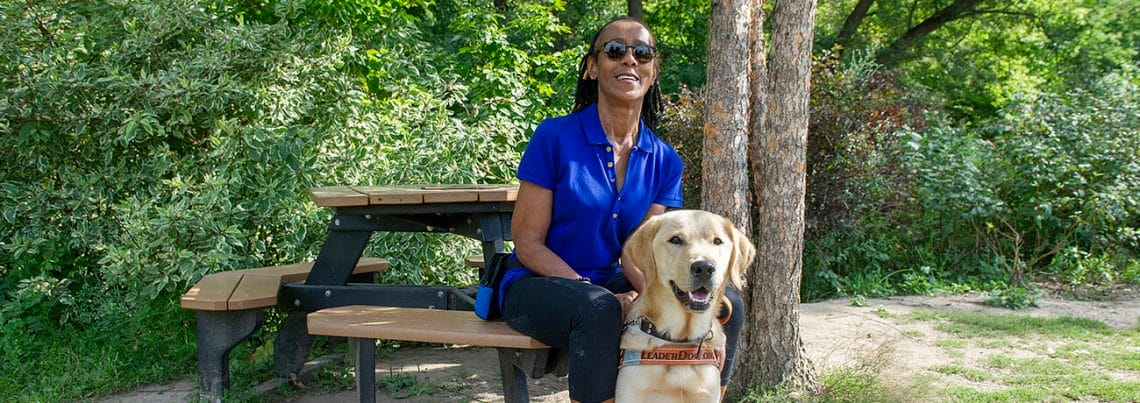 Client Cynthia C LD Duchess sitting smiling park bench trees Smiling woman sitting at picnic table with golden retriever in Leader Dog harness sitting at her feet. Behind them are sunlit trees