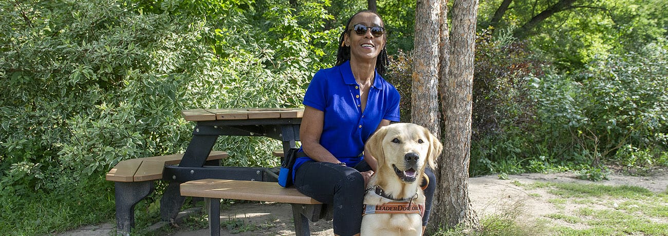 Smiling woman sitting at picnic table with golden retriever in Leader Dog harness sitting at her feet. Behind them are sunlit trees