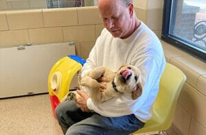 A man in a tan sweatshirt and jeans sits in a yellow plastic chair holding a young yellow lab puppy in his lap. The puppy is licking its nose as the man looks down at it.