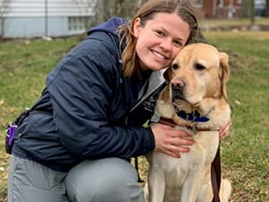 Yellow lab sitting next to smiling woman kneeling on ground with grass behind