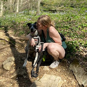 Olivia crouching on dirt path with trees in the background. She is kissing an Australian cattle dog sitting next to her.