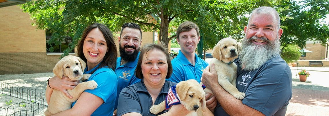 Group of two women and three men all smiling and wearing blue polos with the Leader Dog logo. The three people in front are each holding a yellow lab puppy.