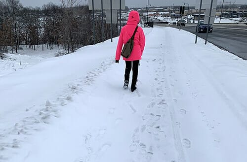 Person facing away from camera walking down side of snowy road with white cane. The person is weraing a bright pink/red coat and dark pants.