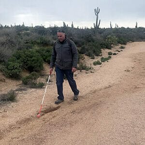 Man in dark jacket and pants walking with white cane in Arizona desert on sandy path