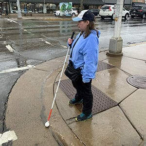 Woman in black pants and blue hoodie stands at curb with cars driving on street in the background. She is holding a white cane in front of her.