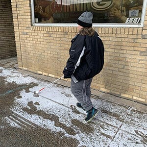 Woman with shoulder-length brown hair walking on snowy sidewalk with a white cane. She is wearing a black jacket and gray sweatpants with tennis shoes.