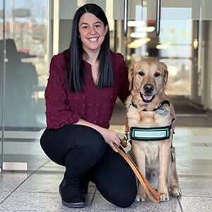 Alyssa, a woman with long dark brown hair, smiles and kneels next to a yellow lab in a green vest that says leaderdog.org on the frost. Alyssa is wearing a maroon blouse and black pants.