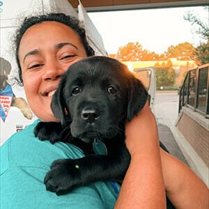 Smiling dark-haired woman holding black lab puppy close to her face.