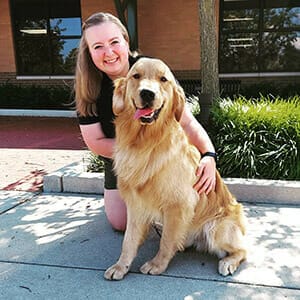 Smiling woman with long brown hair kneels outdoors on sidewalk, hugging golden retriever seated next to her.