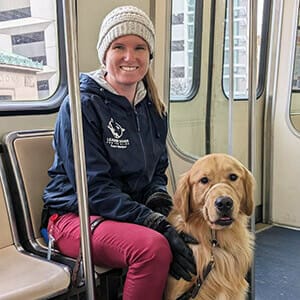 Emily, a woman with blond hair in a ponytail, wearing a blue jacket with the Leader Dog logo, red pants, and a winter hat, smiles while sitting on a trolley next to a golden retriever sitting between her legs.