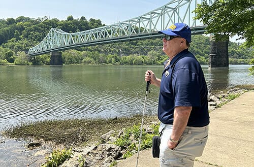 Man standing near a river with a bridge in the background. He is holding a white cane