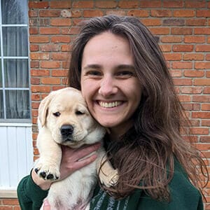 Mara, a woman with long brown hair, smiles while holding a yellow lab puppy close to her face