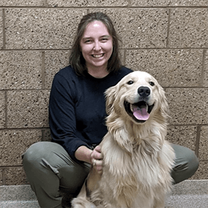 Sarah, a woman with shoulder length brown hair, smiles and crouches next to a golden retriever sitting with its mouth open