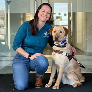 Dark-haired woman smiles while kneeling next to a yellow lab in Leader Dog harness sitting next to her. She is wearing a teal shirt with the Leader Dog logo and jeans.