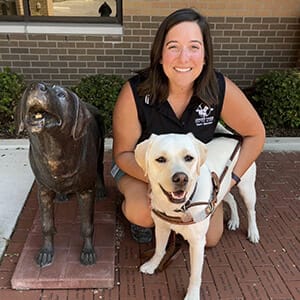 A smiling woman with brown hair kneels next to a yellow lab in a Leader Dog harness. Lauren is wearing a black sleeveless shirt with the Leader Dog logo.