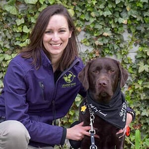Meredith smiles while kneeling next to a chocolate lab in a black bandanna. Meredith has shoulder-length brown hair and is wearing a purple jacket with the Leader Dog logo.