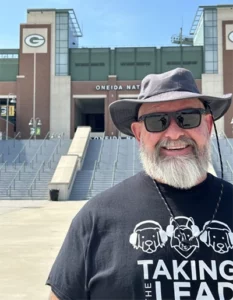 Man in sunglasses, floppy hat and a t-shirt smiling at the camera with Lambeau Field's entrance visible in the background.