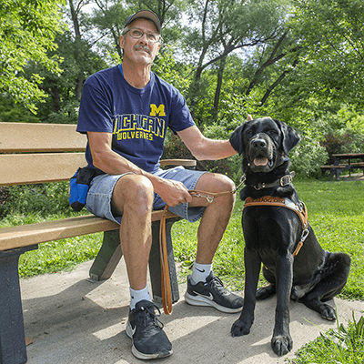 Client Wayne Sanford LD Otto sitting park bench (1) Man wearing a University of Michigan t-shirt and jean shorts sits on a park bench next to a black lab in Leader Dog harness that's sitting on the ground. There are trees and grass in the background