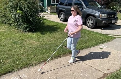 Woman walking down sidewalk with grass and a house behind her. She is walking with a white cane.