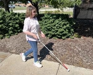 Woman walking with white cane down sunny sidewalk with wood chips and bushes in the background.