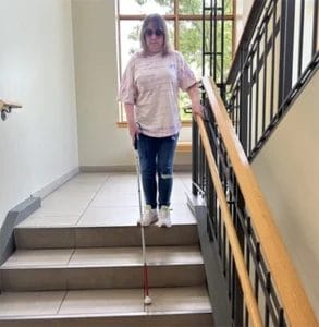 Woman walking toward the camera down partially open stairs using a white cane. 