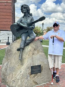 Smiling man in sunglasses, t-shirt, and shorts standing next to statue of Dolly Parton perched on a large rock with a guitar in her hands