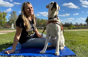 Woman sitting outdoors on blue blanket on grass smiling at a yellow lab in Leader Dog harness sitting next to her