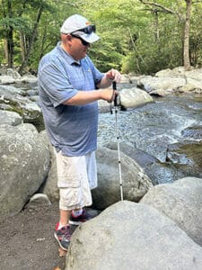 Man with white cane standing near large rocks at the edge of a creek with woods in the background