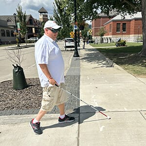 Man in shorts, t-shirt, and ballcap walking on a sunny sidewalk with white cane