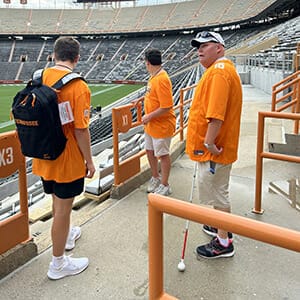 Three men in bright orange shirts standing inside a stadium in the bleachers