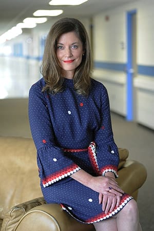 Melissa sits on the edge of a chair, smiling at the camera. She is wearing a blue dress with red and white accents