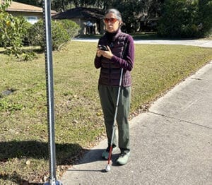 Woman standing outdoors on a sidewalk near a road sign. She is holding a white cane