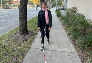 Smiling woman walking down a sidewalk toward the camera with a white cane. There is grass on either side of the sidewalk and some trees lining the sidewalk