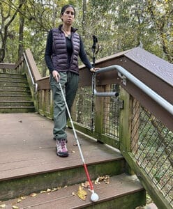 Woman standing at a landing on outdoor wooden steps with trees in the background. She is holding a white cane