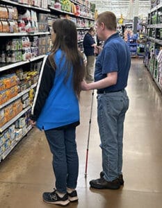 Man with white cane in one hand stands near a woman while holding her elbow as she acts as a sighted guide. They are facing a shelf of products in a department store