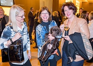 Three women stand smiling with two looking at the camera and the third looking at a chocolate lab puppy being held by the woman in the middle. A crowd of people is visible in the background