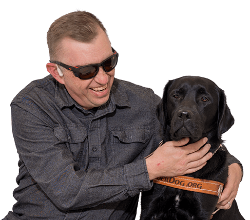 Man with wide smile on his face as though he's mid-laugh with his arms around a black lab sitting next to him. The dog has a brown Leader Dog harness on. The man is wearing sunglasses and a charcoal button-up shirt