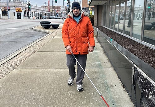 Man walking toward camera on a sidewalk with a white cane. He's wearing an orange coat and black winter hat.